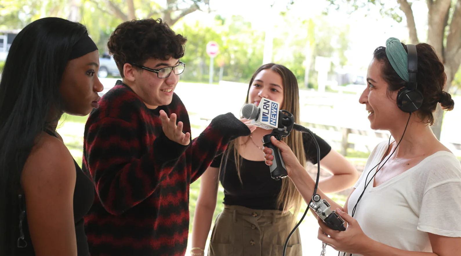 A young man speaks into a microphone held by a woman wearing headphones while two other young people stand nearby outdoors.